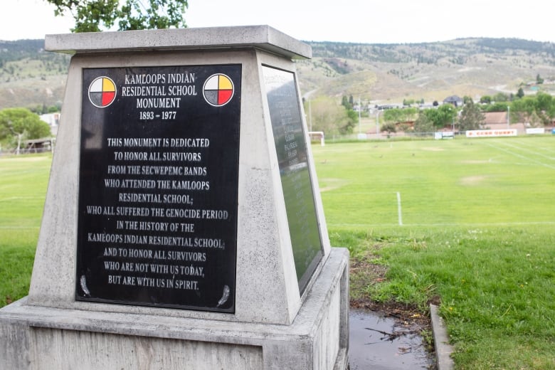 A stone monument stands before a green field. A black plaque dedicates the monument to survivors of the Kamloops Residential School. 