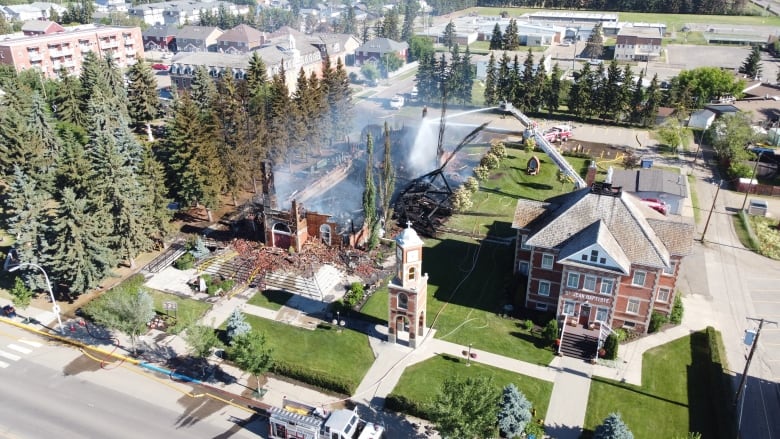 An aerial image shows a firefighter on a ladder truck directing spray from a fire hose at the smouldering remains of a church building that is surrounded by pine trees and other buildings. 