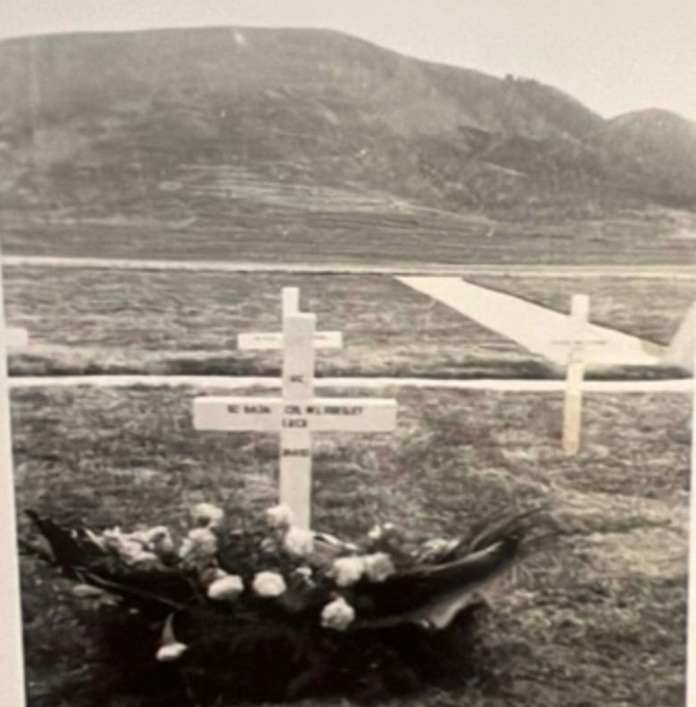 A black and white photograph of a grave marked with a cross and flowers. 