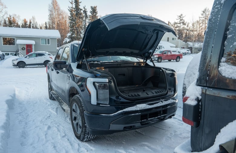 A truck parked in a snowy driveway, with a residential neighborhood visible in the background. It's hood is raised, revealing storage space where the combustion engine of gas-powered car would go. 