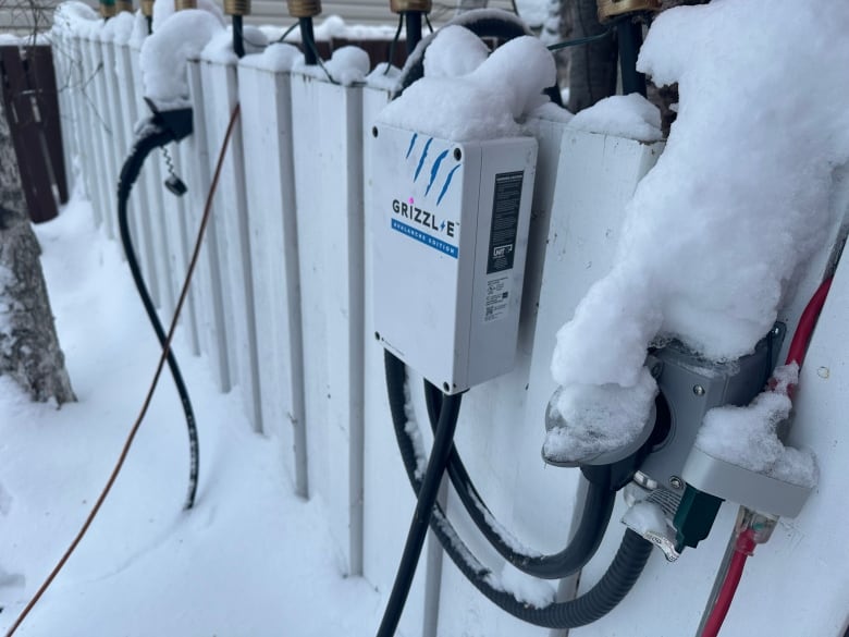 A white box installed on a white fence that's covered in snow. Its outlet and charging nozzle are also affixed to the fence.