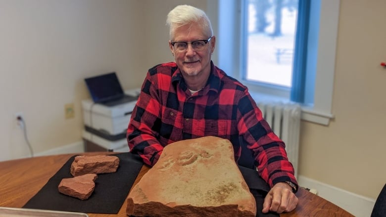 A man in a red plaid shirt sits in front of a fossil -- the imprints of tracks on a large slab of red clay rock.