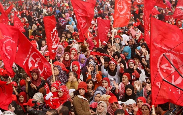 A crowd of women, some wearing head scarves, hold red flags. 