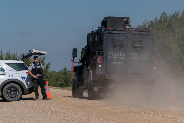 A large black armoured vehicle drives on a dirt road past an RCMP SUV with an officer standing outside.