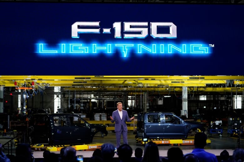 A man in a blue suit speaks to a crowd of people in front of two electric vehicles at various points in the assembly process. A neon sign above reads F-150 Lightning 