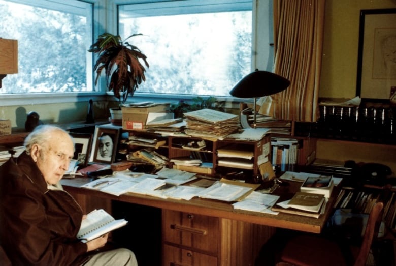 An elderly man with thin white hair is in the bottom left corner of the photo. He is in a study room filled with papers and books.