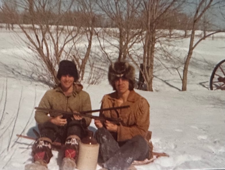 Two smiling men sit in the snow while holding guns. 