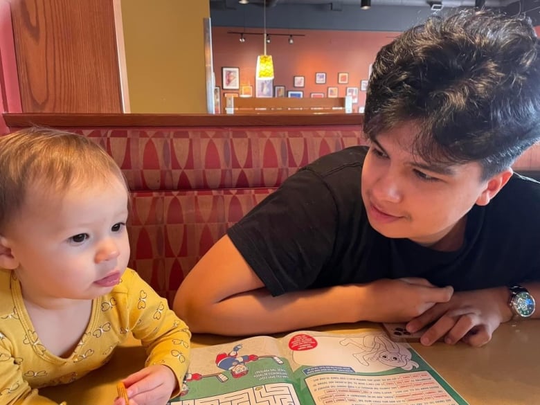 A young man and a toddler sit together in a booth at a restaurant with a colouring book on the table in front of them.