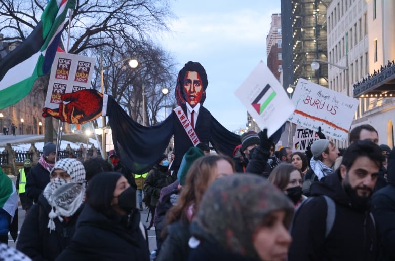 People protest on a city street in winter. One person has a caricature of a politician wearing a sash that says 'war crime.'