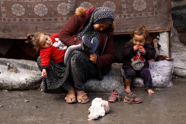 A woman sits on a white sack, with one child on her lap and another sitting next to her in Rafah.