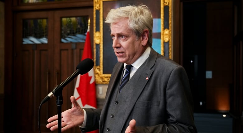 Charlie Angus stands at a microphone, holding his open hands to his side, wearing a three-piece suit. 