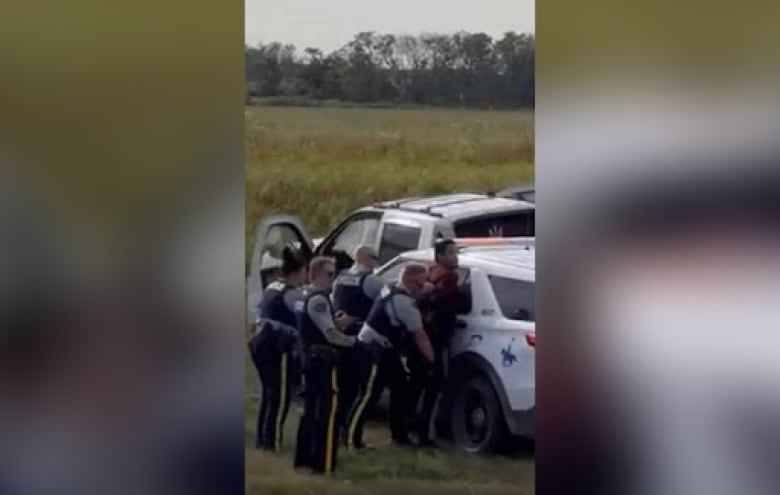 A blurry cellphone photo shows multiple police officers standing behind a man who is pressed up against an RCMP vehicle and has his hands behind his back.