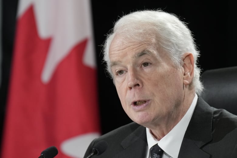 Man with concerned expression, white hair and wearing suit and tie speaks into a microphone in front of Canada flag