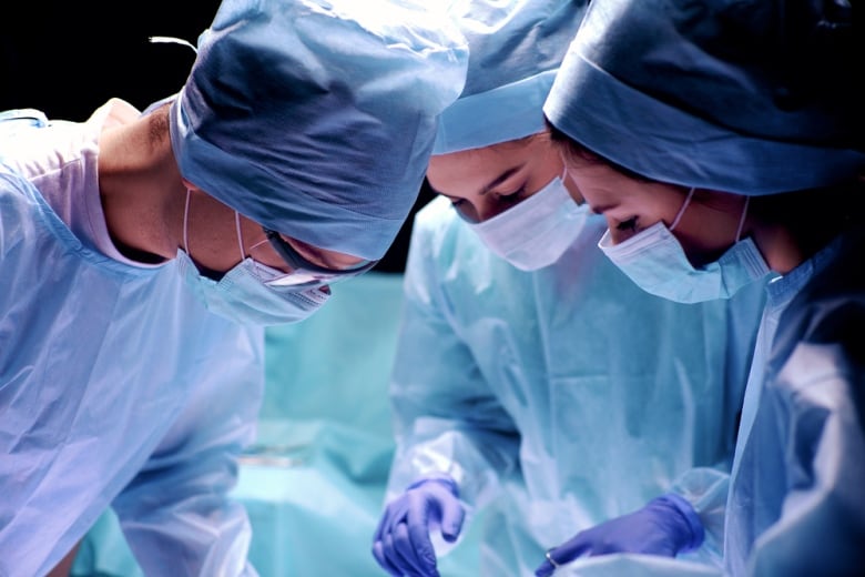 Three surgeons wearing scrubs are pictured looking down at a patient in an operating room.