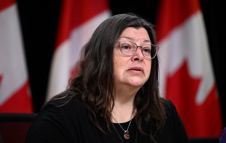 A woman with dark hair and glasses, with Canadian flags displayed in the background.