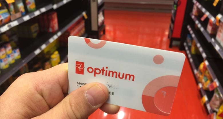 A loyalty card with the words PC Optimum on the front is seen close-up in a person's hand. The aisle of a retail store is seen in the background.