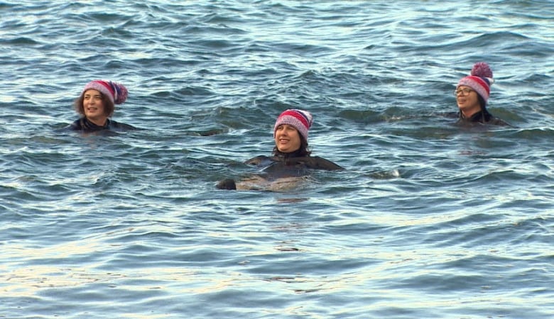 Three women swimming in the ocean while wearing winter hats. 