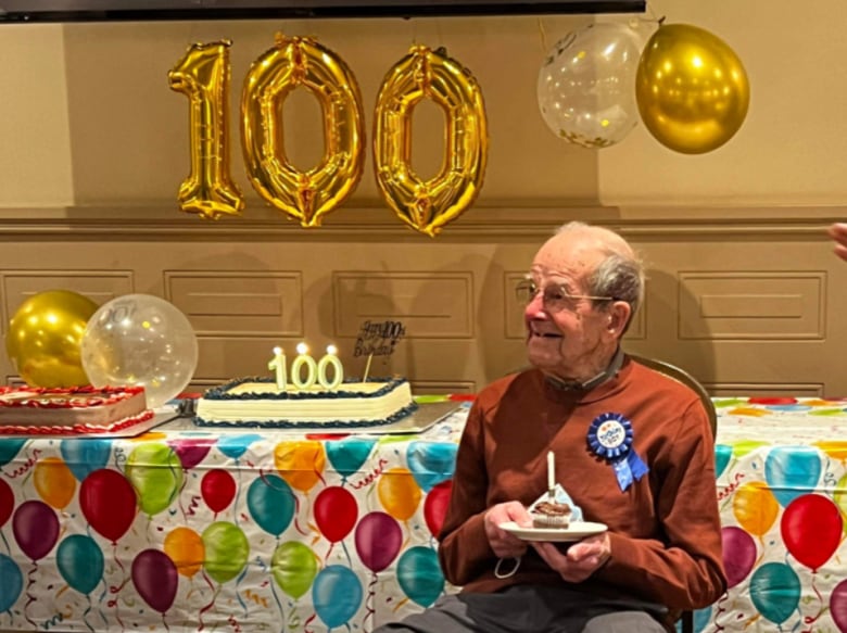 A man sit sin a chair olding a cupcake with a candle on it. Behind him are birthday balloons and a birthday cake with a numbered birthday candle. 