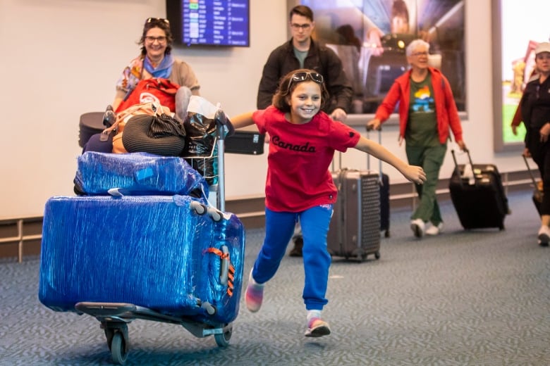 A smiling girl runs in a red Canada t-shirt beside blue luggage at YVR.