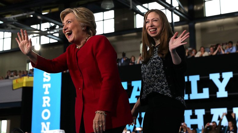 Hillary and Chelsea Clinton smiling and waving on stage as audience claps and takes photos