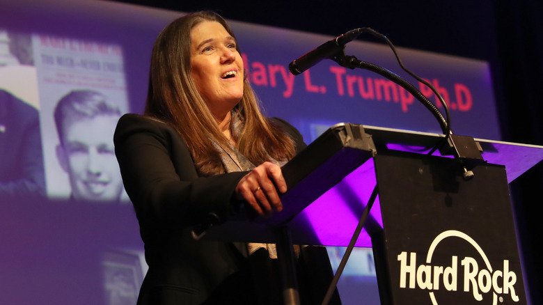 Mary Trump speaking at an event with an image of Donald Trump in the background