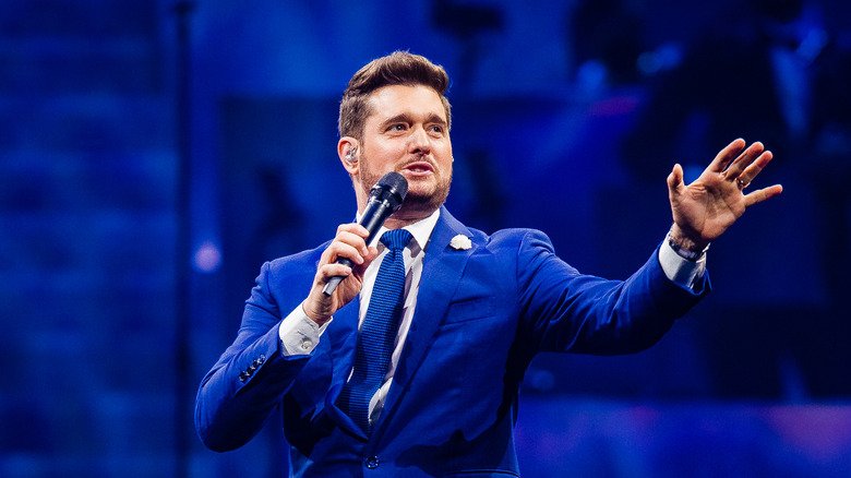 Michael Bublé wearing a blue suit, blue tie, and white flower lapel singing into a microphone