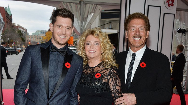 Michael Bublé wearing a dark blue grey suit with his parents all wearing a red flowered lapel