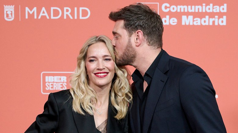 Michael Bublé wearing a black suit and shirt kissing his wife Luisana Lopilato on the head as she smiles