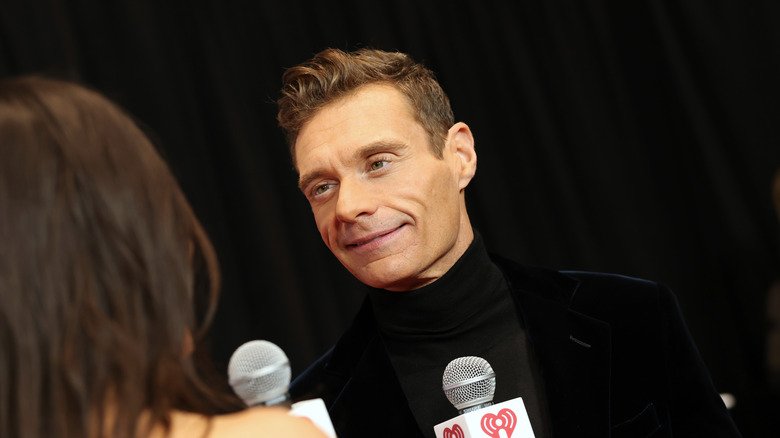 New Spotlight, Old Worries: Ryan Seacrest’s Dramatic Look Steals the Moment 110 Ryan Seacrest looking at a host while holding a microphone in a black turtleneck with a matching velvet blazer