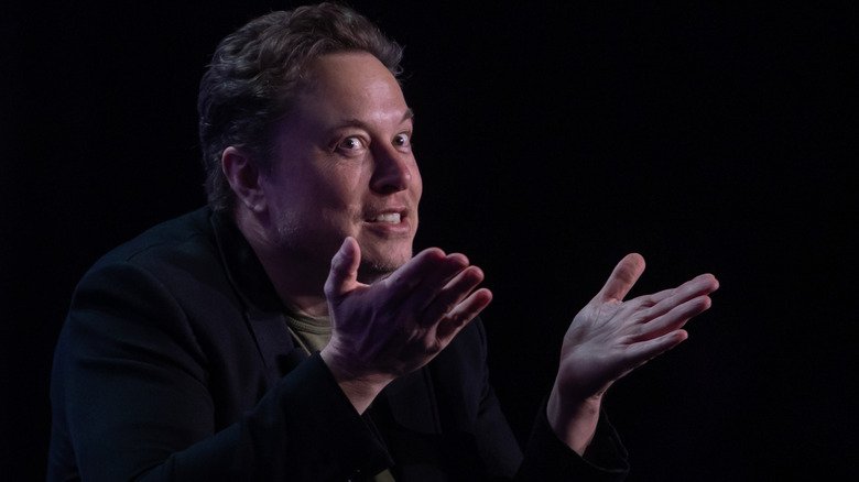 Elon Musk speaking animatedly on stage at a conference in a black blazer