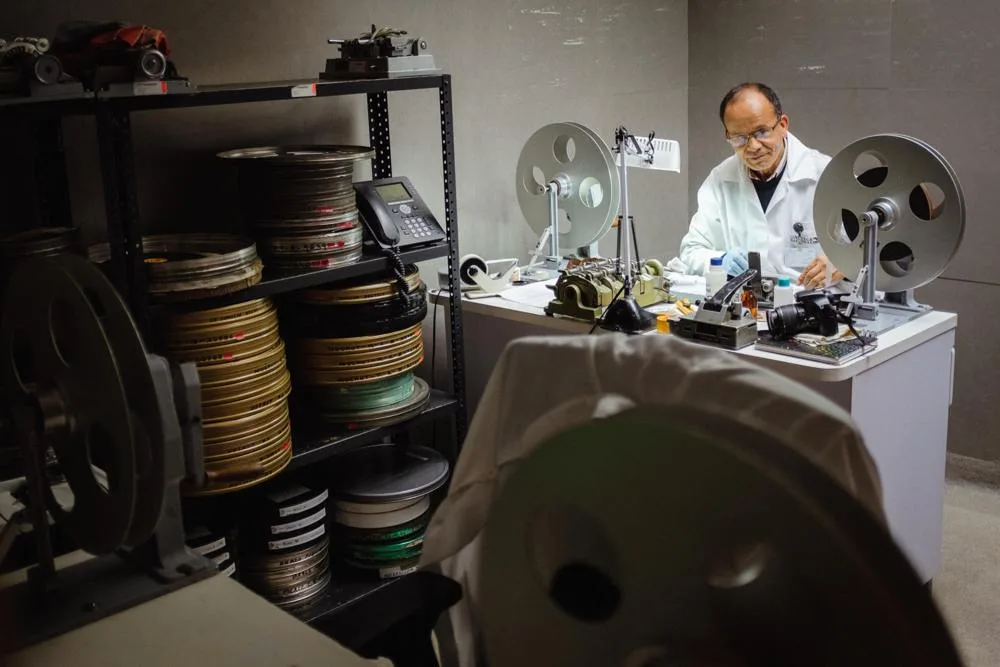 A film-restoration worker handles material at a workbench with reels and film canisters.