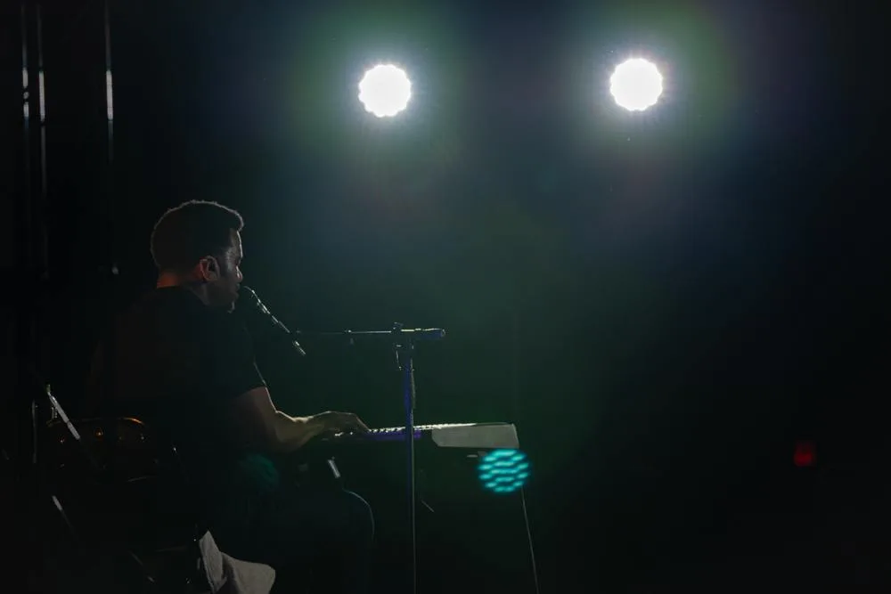 Craig Robinson is seen from the side playing a keyboard on a dark stage at Camp Casey.