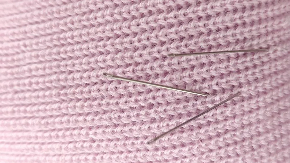 A close-up of hand-sewing needles resting in pale pink knitted fabric